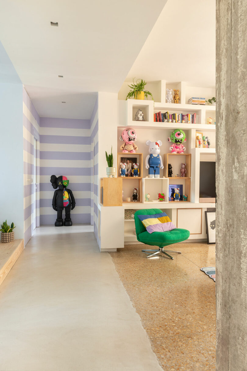 Modern living room with striped walls, colorful art toys, a green swivel chair, and built-in shelves with books and plants.