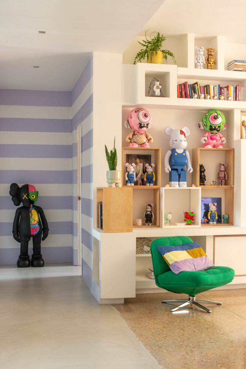 Room with striped wall, large figurines on display shelves, books, a green chair with a colorful cushion, and a small plant.