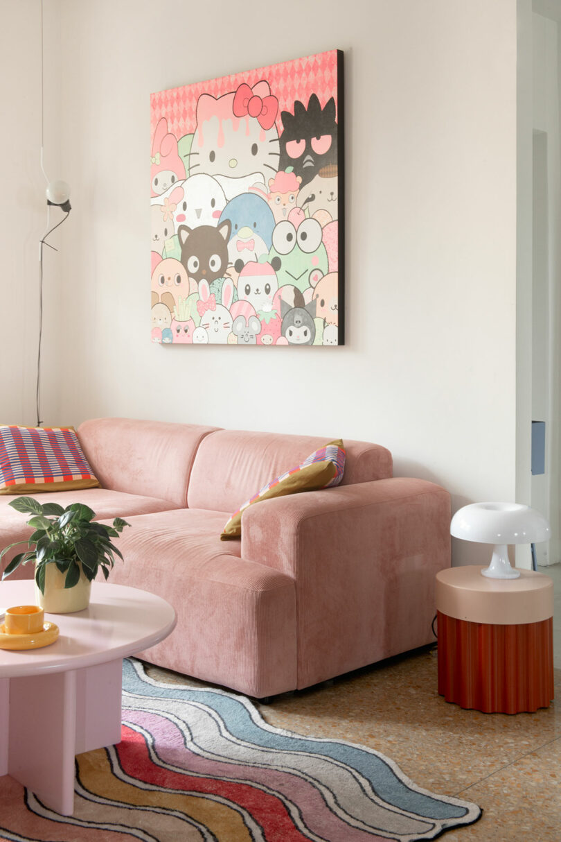 A living room featuring a pink sofa, a wavy-patterned rug, a round pink coffee table with plants and cups, and a wall art piece with various cartoon characters.