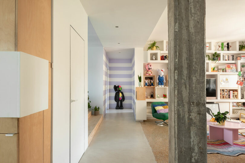 A modern living space with a hallway featuring a striped wall, art figures on display, and colorful furniture including a round chair and a low table.