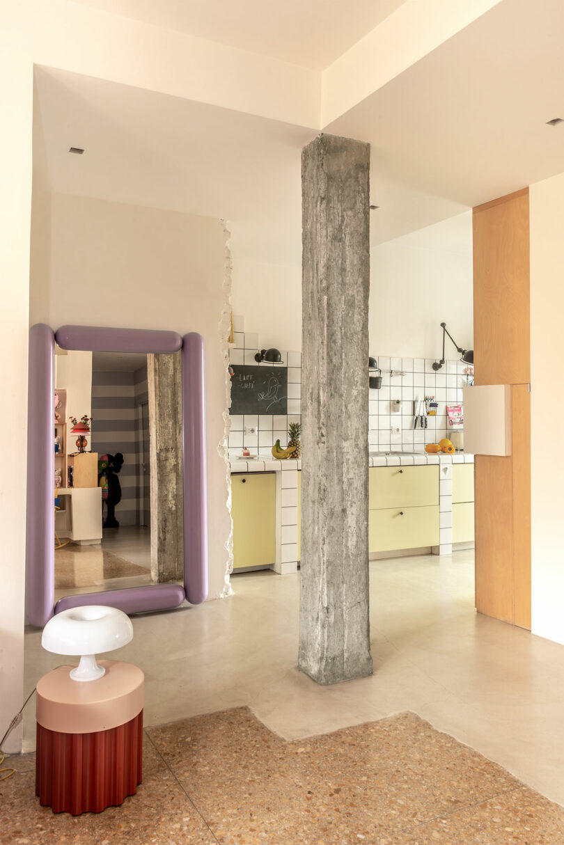 Interior view of a modern kitchen with light yellow cabinets and white tiled walls. A concrete pillar stands in the center. A purple framed mirror and a small lamp on a table are visible.