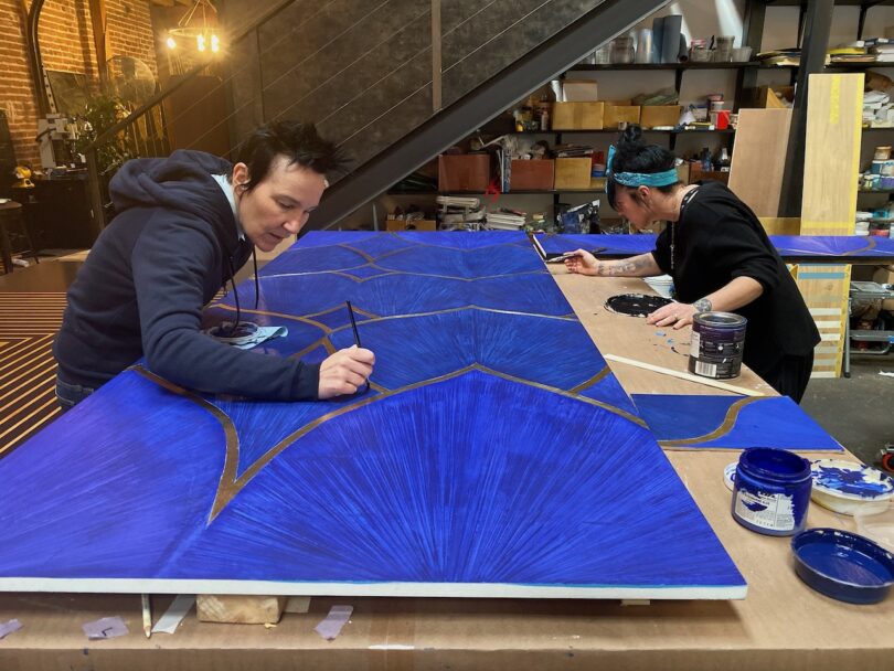 Two people are painting large blue geometric designs on wooden panels in an art studio