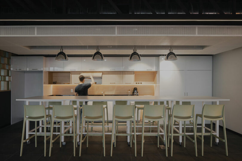 A person standing in a modern kitchen with white cabinets, a long island with chairs, pendant lights, and a built-in coffee machine.