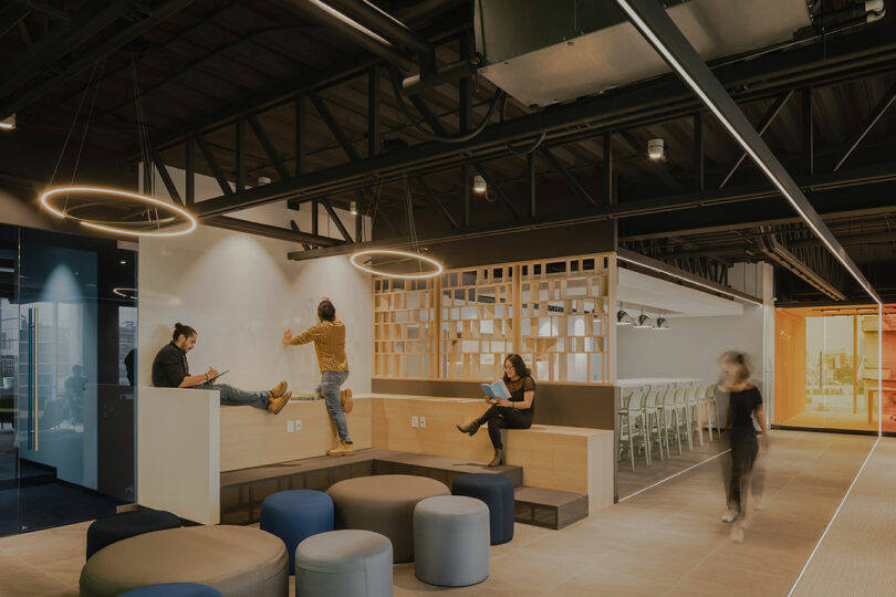 Modern office space with people using laptops, sitting on tiered seating. Circular lights hang from the ceiling, and an open area with seating and a bar is visible in the background.