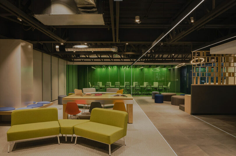 Modern office space with green and beige sofas, a wooden table with colorful chairs, and a green curtain in the background. Overhead lighting is visible, with industrial ceiling design.