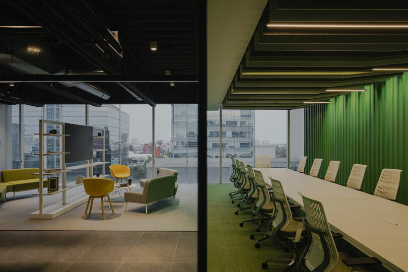 Modern office space with a glass-walled seating area on the left, featuring chairs and a TV, and a conference room on the right with a long table and white chairs.