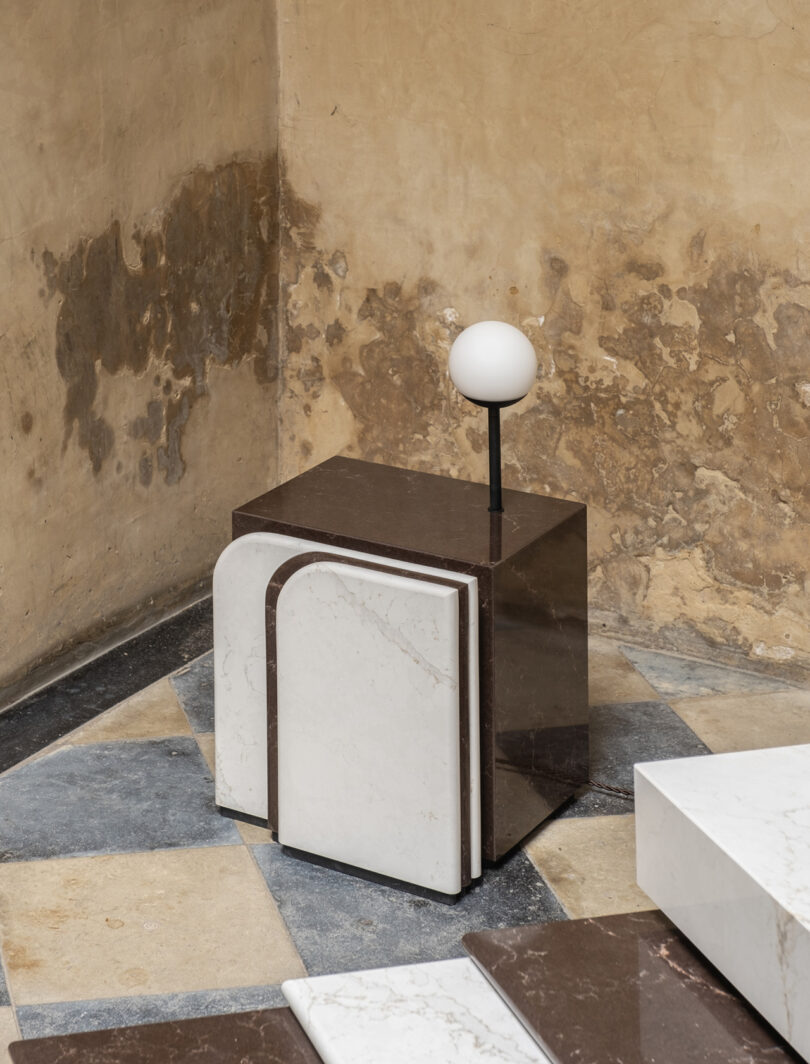 A geometric sculpture with a marble base and a spherical white top is placed in a corner with textured beige walls.