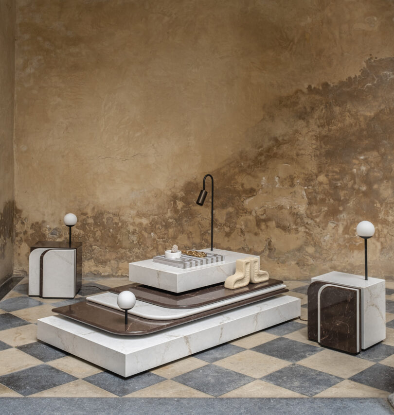 Artistic installation featuring geometric shapes and minimalist decor in a rustic setting, with a chessboard on a layered marble platform and modern lamp accents.