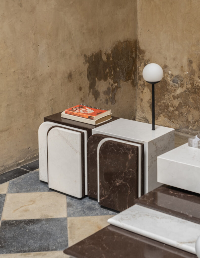 Two geometric marble tables, one white and one brown, with a book and a small lamp on top. They are set on a checkered floor in a room with textured walls.