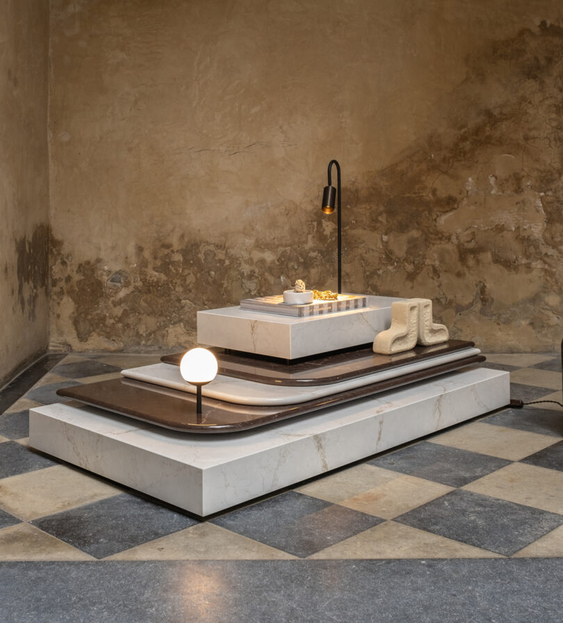 Art installation with sculpted blocks, curved layers, a small table lamp, and textured boots on a checkered floor in front of a worn wall.