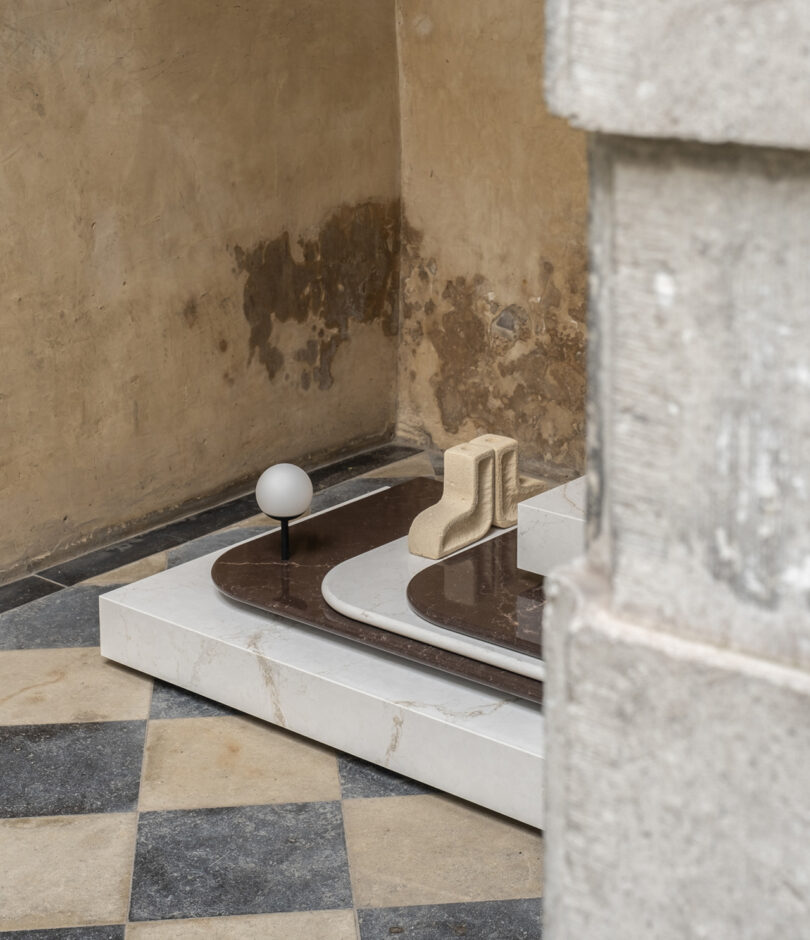 Abstract architectural model with geometric shapes and varying textures on a checkered floor, set against a worn wall.