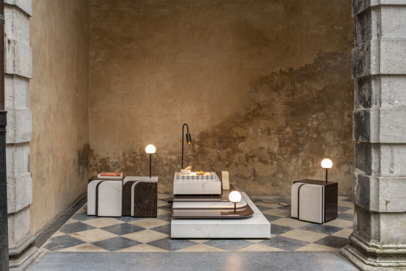 Modern minimalist room with marble furniture, a low table with books and lamps, and a checkered floor, set against a textured stone wall.