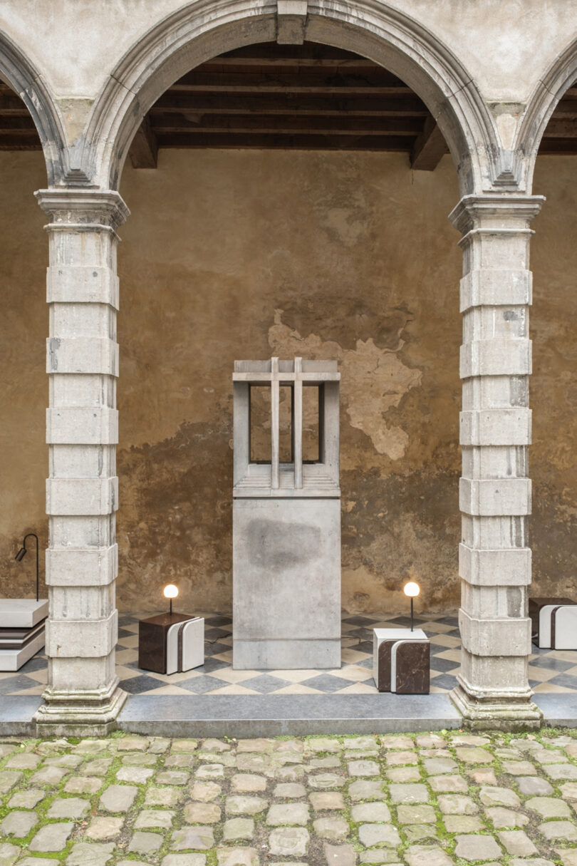 A stone sculpture stands within an arched stone structure, flanked by two small lamps and geometric blocks on a checkered floor.