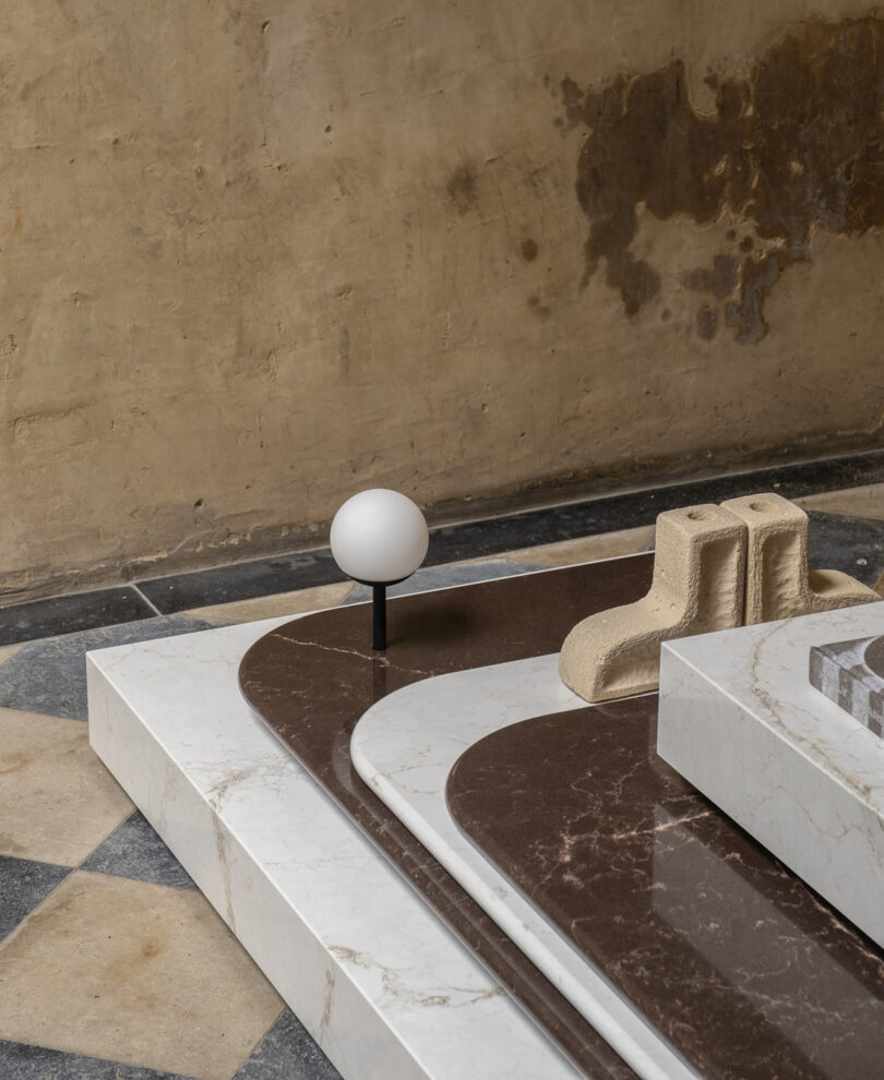 A geometric installation with layered marble and stone slabs, featuring a small spherical light and a textured abstract sculpture, set against a worn wall backdrop.