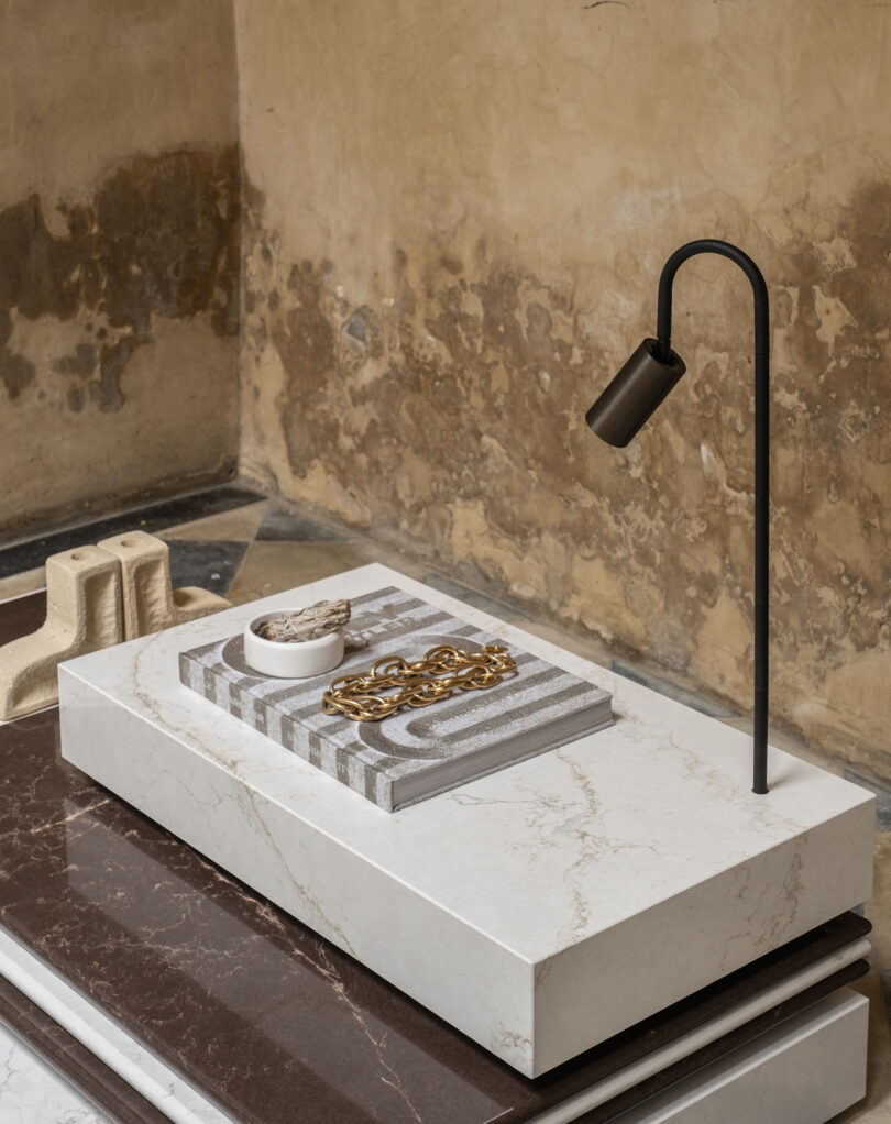 Marble table with a modern lamp, two books, a chain, and a small bowl against a textured wall background.