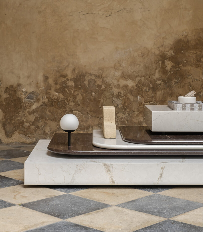 A minimalist geometric marble display features stacked rectangular slabs, a small sphere on a stand, and a sponge, set against a weathered wall and checkered floor.