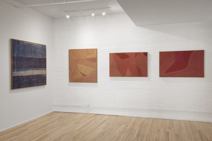 Four abstract paintings hang on a white brick wall in a gallery with wooden flooring and overhead lights.