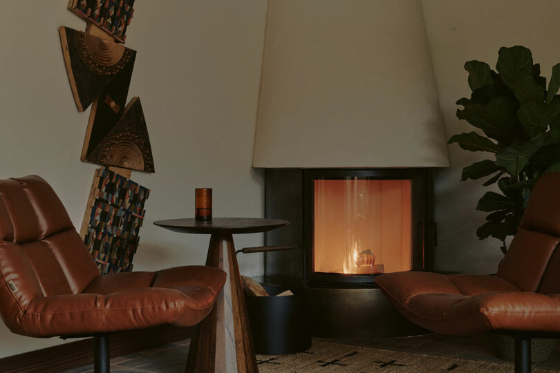 A cozy living room with two brown leather chairs facing a modern fireplace. A round wooden table holds a candle. Geometric wall art and a potted plant are in the background.