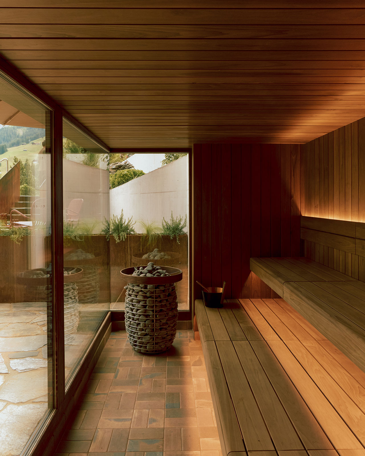 A modern wooden sauna room with large glass windows, a bench, and a stone heater. Outside, there's a view of a patio and greenery.