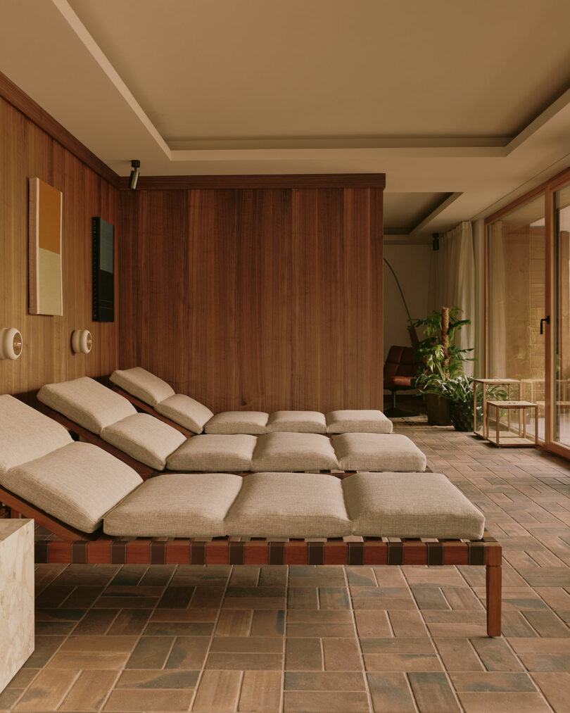 Lounge chairs with beige cushions line a wooden wall in a minimalistic room with brick flooring and potted plants by a large window.