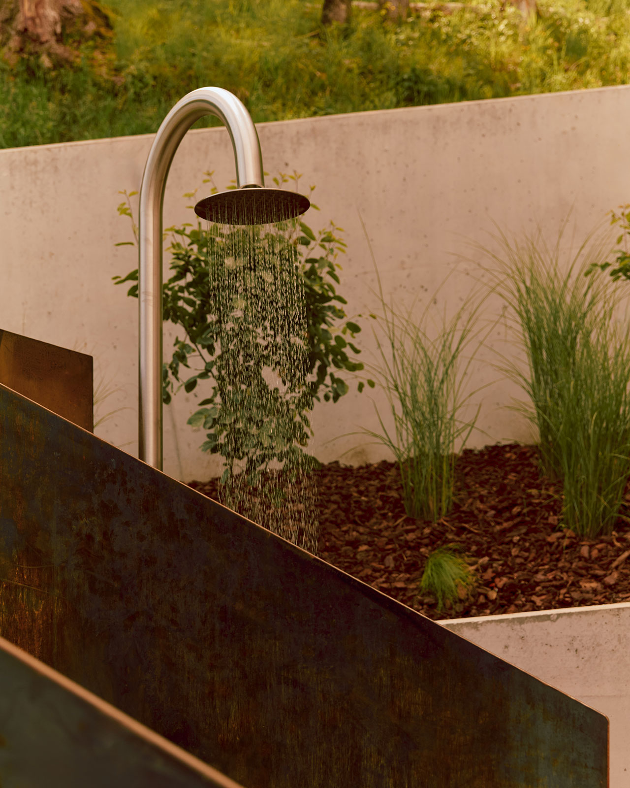 An outdoor shower with running water is set against a concrete wall, with greenery and mulch in the background.