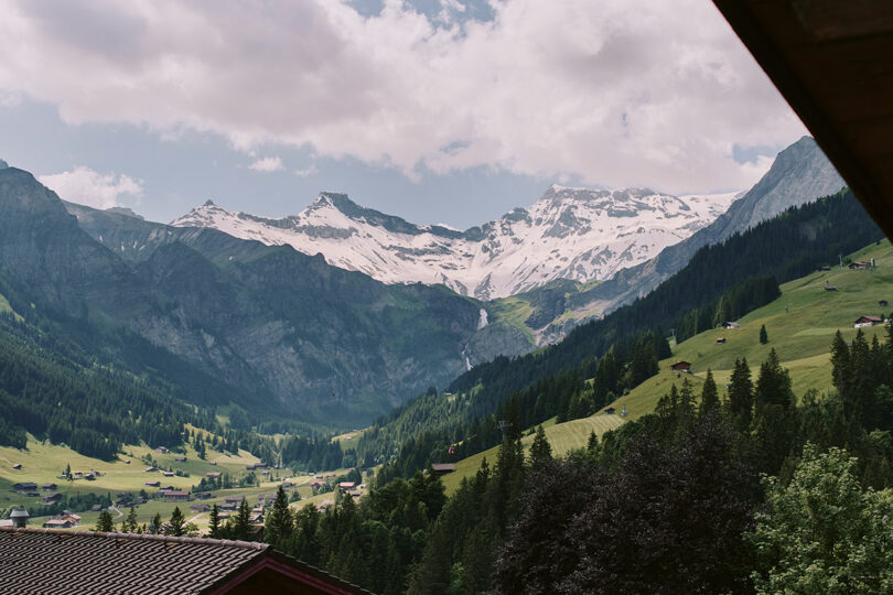 Mountain landscape with snow-capped peaks, green valleys, scattered houses, and a waterfall under a partly cloudy sky.