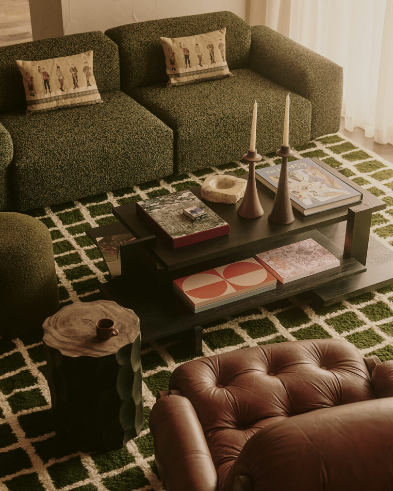 A cozy living room with two green sofas and a leather armchair. A dark coffee table with candles, books, and decor sits on a patterned rug.