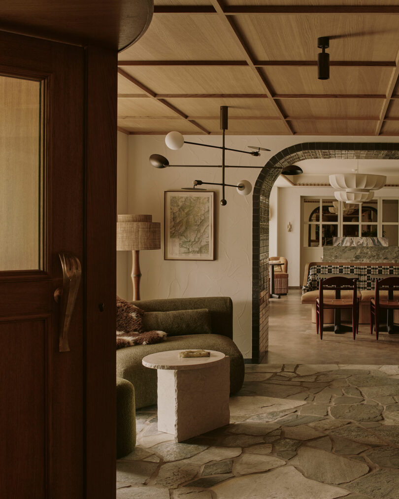 A cozy interior featuring a green couch, stone coffee table, and wooden ceiling. A modern chandelier hangs above. A dining area is visible through an arched doorway.