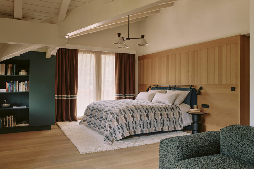 Cozy bedroom with a double bed, geometric bedding, a wood accent wall, dark curtains, bookshelves, and a green chair. Natural light filters through a large window.