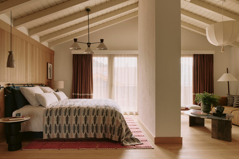 A cozy bedroom with a large bed, patterned bedspread, wood-paneled walls, and vaulted ceiling. Curtains cover wide windows. A seating area with a round table and sofa is visible.