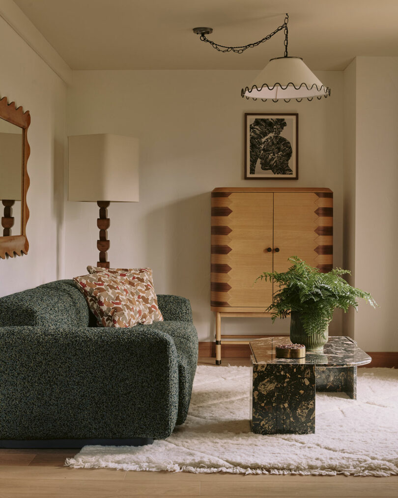 Cozy living room with a green sofa, patterned cushion, tall lamp, wooden cabinet, framed art, marble coffee table, potted fern, and a shaggy white rug.