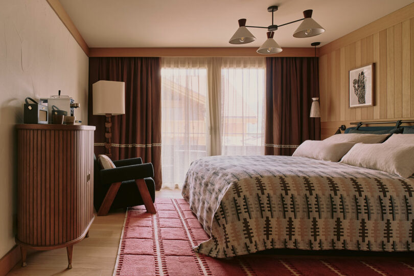 Cozy bedroom with a large bed, patterned bedding, dark curtains, wooden accents, and a retro-style armchair. A ceiling light fixture, framed artwork, and a side cabinet complete the decor.