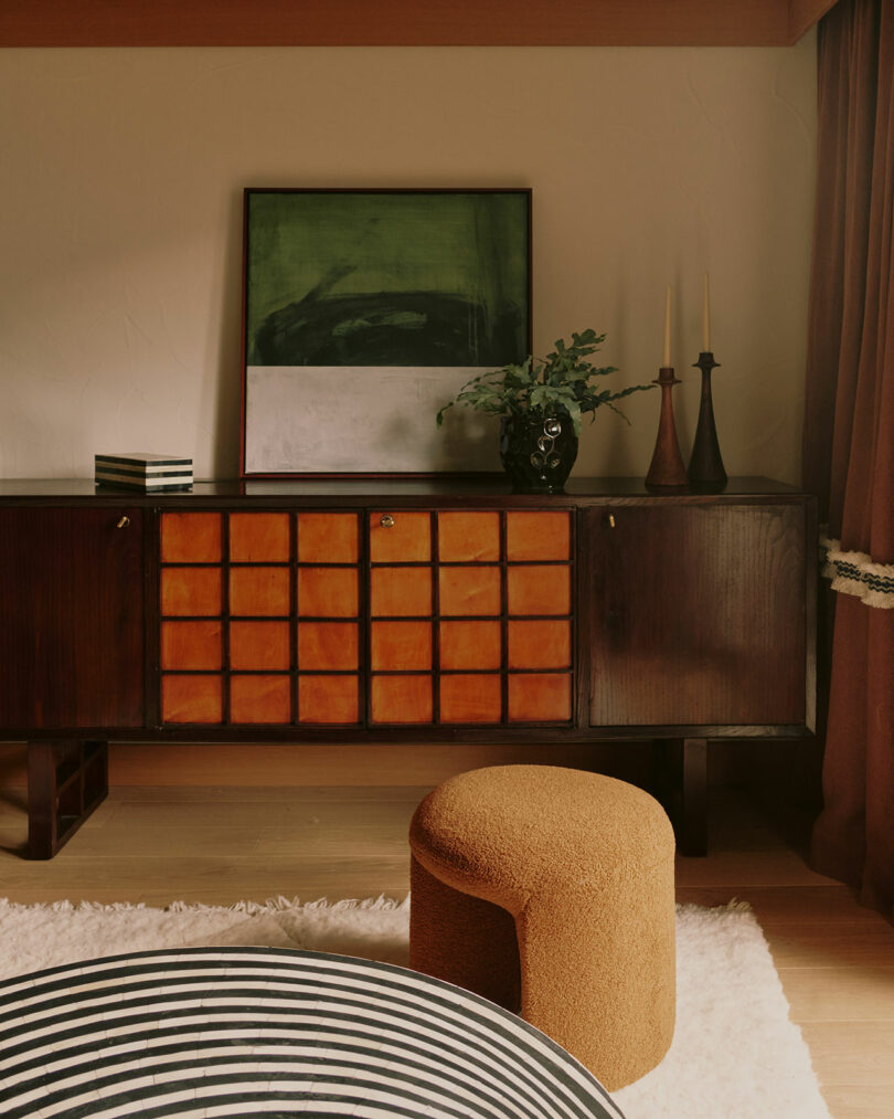 Mid-century modern room with a wooden sideboard, abstract art, two tall candles, a green plant, brown ottoman, and a black-and-white striped cushion on a beige rug.