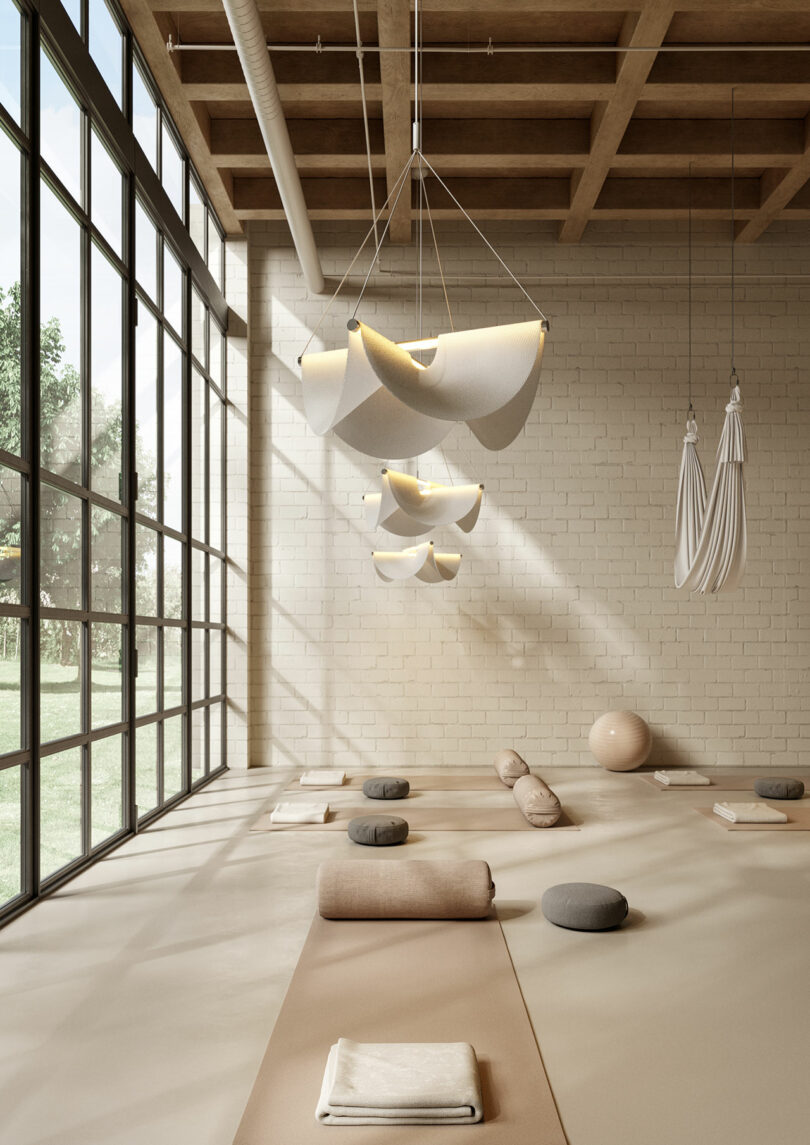 A sunlit yoga studio with large windows features neatly arranged yoga mats and cushions. Soft lighting fixtures drape light gracefully under a wooden ceiling, creating an inviting ambiance.