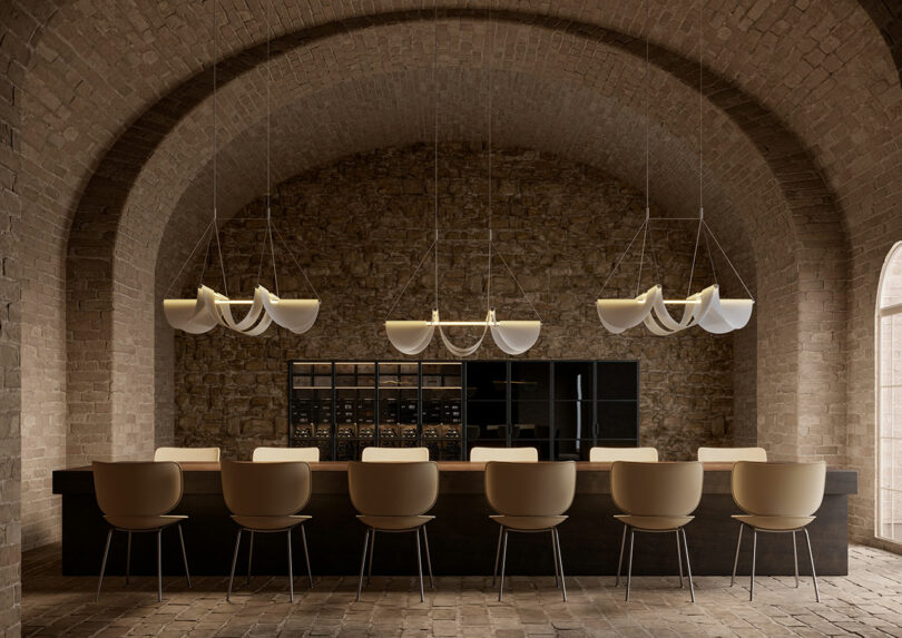 A modern bar with a long counter features beige stools and drape lights hanging elegantly against a brick wall, all under an arched ceiling.