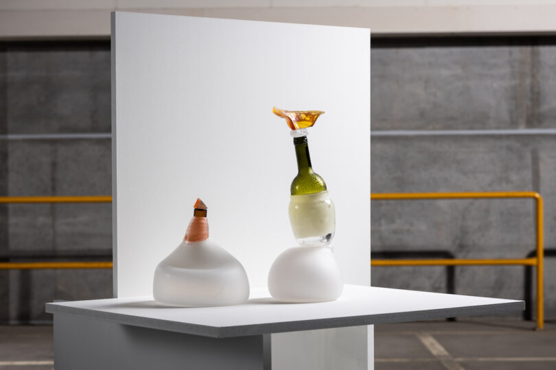 Two abstract sculptures on a white platform: one with an upturned beer bottle, frothy liquid, and wax; the other with a wax-covered neck and bottle cap, in front of a white backdrop