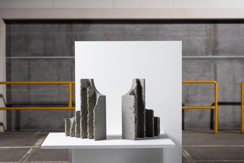 Abstract concrete sculptures on a white pedestal, set against an industrial-style background with yellow railings.