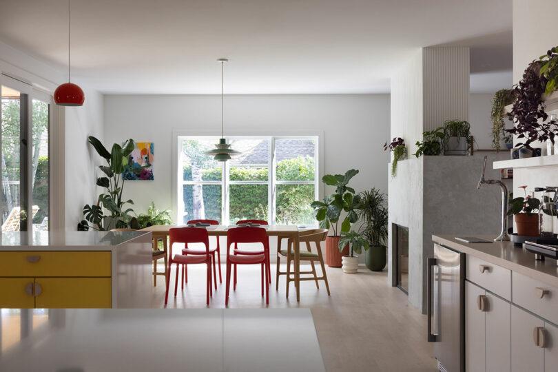 Bright open kitchen and dining area with large windows, red chairs around a table, plants, and modern decor.