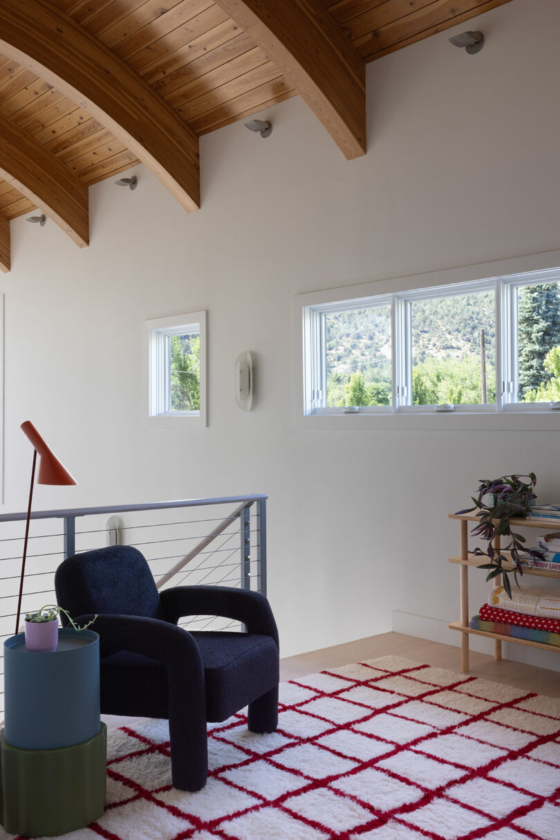 A modern room with a dark armchair, a small side table, and a checkered rug. Three windows offer a view of trees and mountains. A bookshelf with plants stands near the corner.