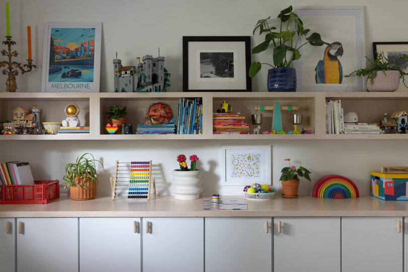 A shelf with books, toys, plants, and framed photos. A cabinet with closed doors is below the shelf. The room is well-lit, with a colorful and organized arrangement.