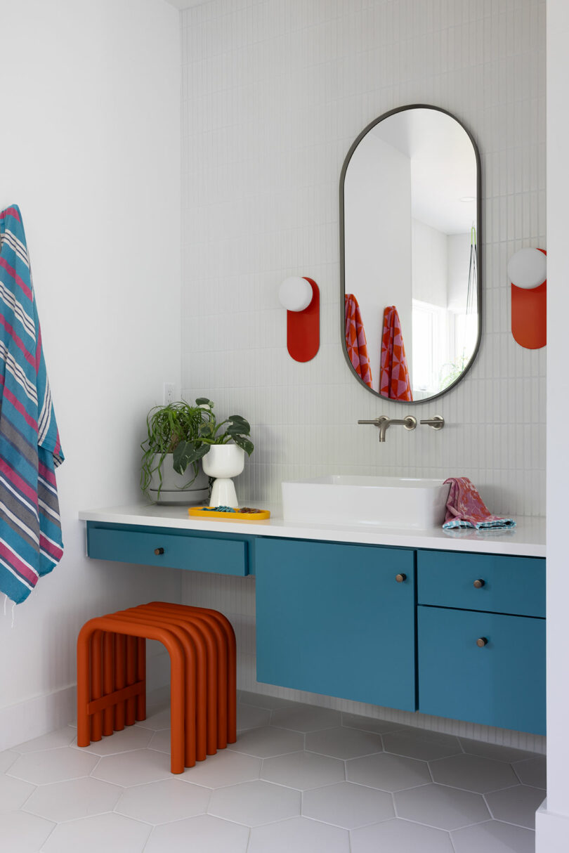 Modern bathroom with a blue vanity, oval mirror, and wall-mounted faucet. Red towel accents and an orange stool add color. A plant sits beside the sink.