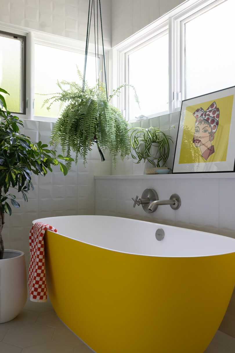 A modern bathroom with a vibrant yellow bathtub, potted plants, a patterned towel, and framed artwork on the ledge.