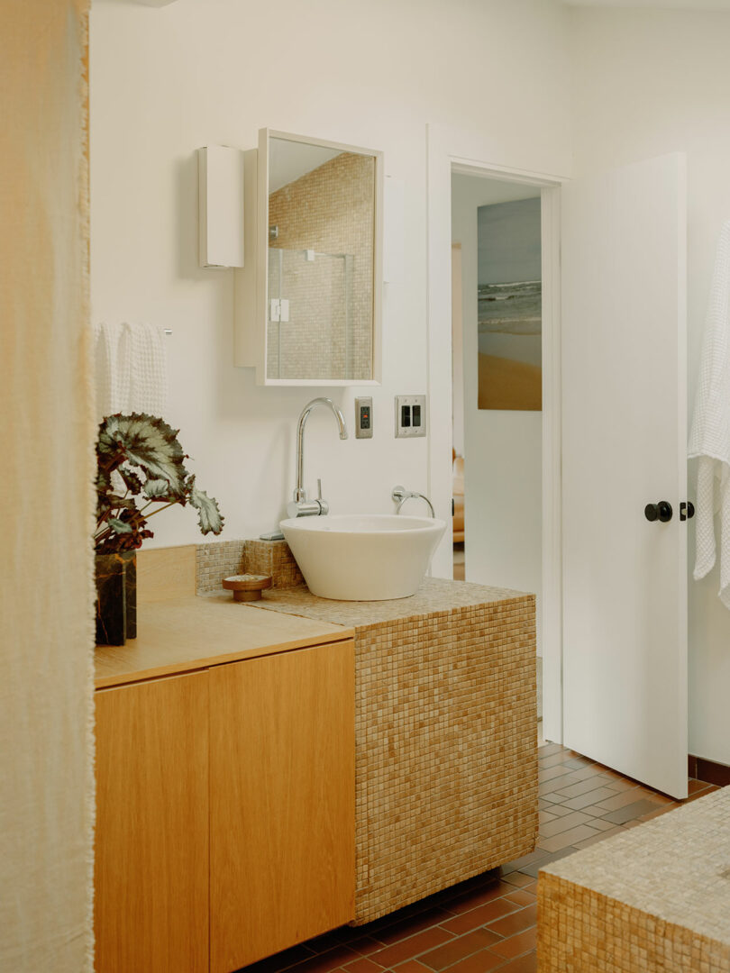 Modern bathroom with a round white sink, wooden cabinetry, and a wall-mounted mirror. Tiled surfaces in earth tones and a potted plant decorate the space. Door slightly ajar in the background.