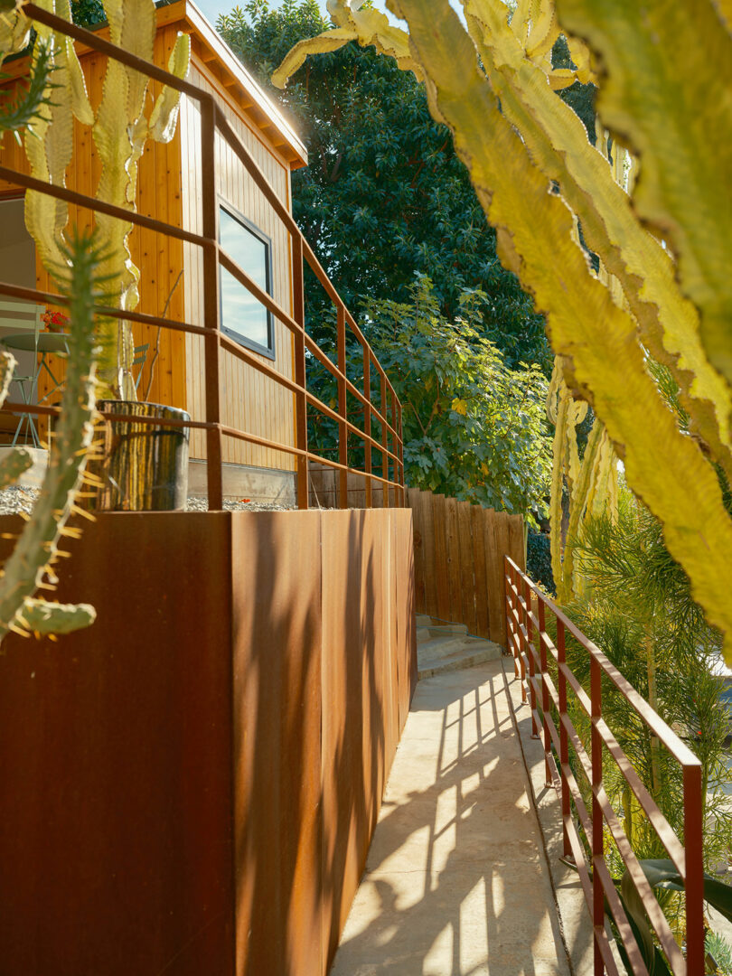 A narrow outdoor path with metal railings runs alongside a wooden building. Tall cacti and trees border the walkway, creating a sunlit, natural setting.