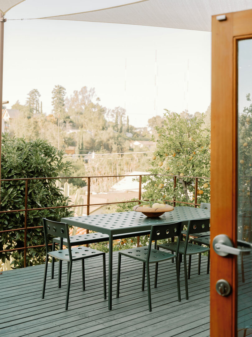 Outdoor deck with a metal dining table, four chairs, and a bowl on top. Surrounded by greenery, enclosed by railing, with a view of the landscape. Wooden door in the foreground.