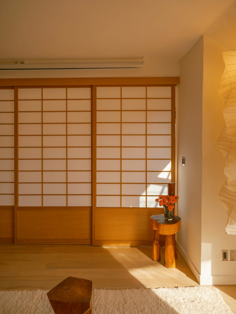 A room with wooden shoji doors, a small wooden table holding a vase of orange flowers, and a textured white lamp on the side. Sunlight casts shadows on the door.