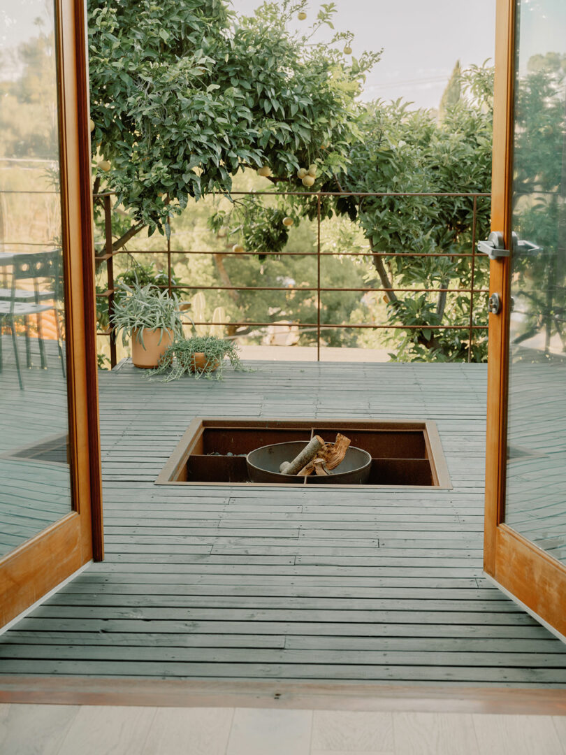 Open patio doors leading to a deck with a central fire pit, surrounded by lush greenery and trees in the background. A potted plant is placed on the left side of the deck.