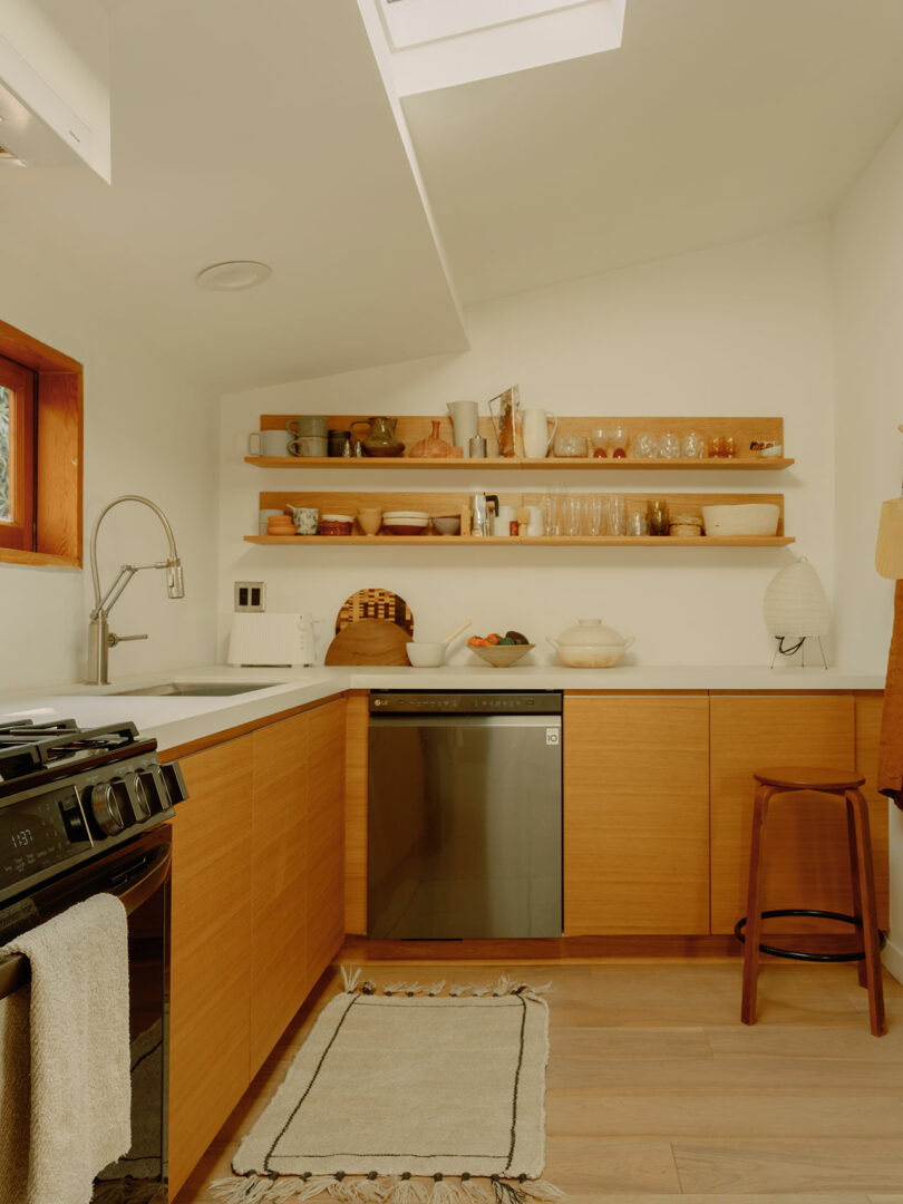 Minimalist kitchen with wooden cabinets, open shelves holding dishes, a stove, and a dishwasher. A skylight brightens the space, and a small rug lies on the wooden floor.