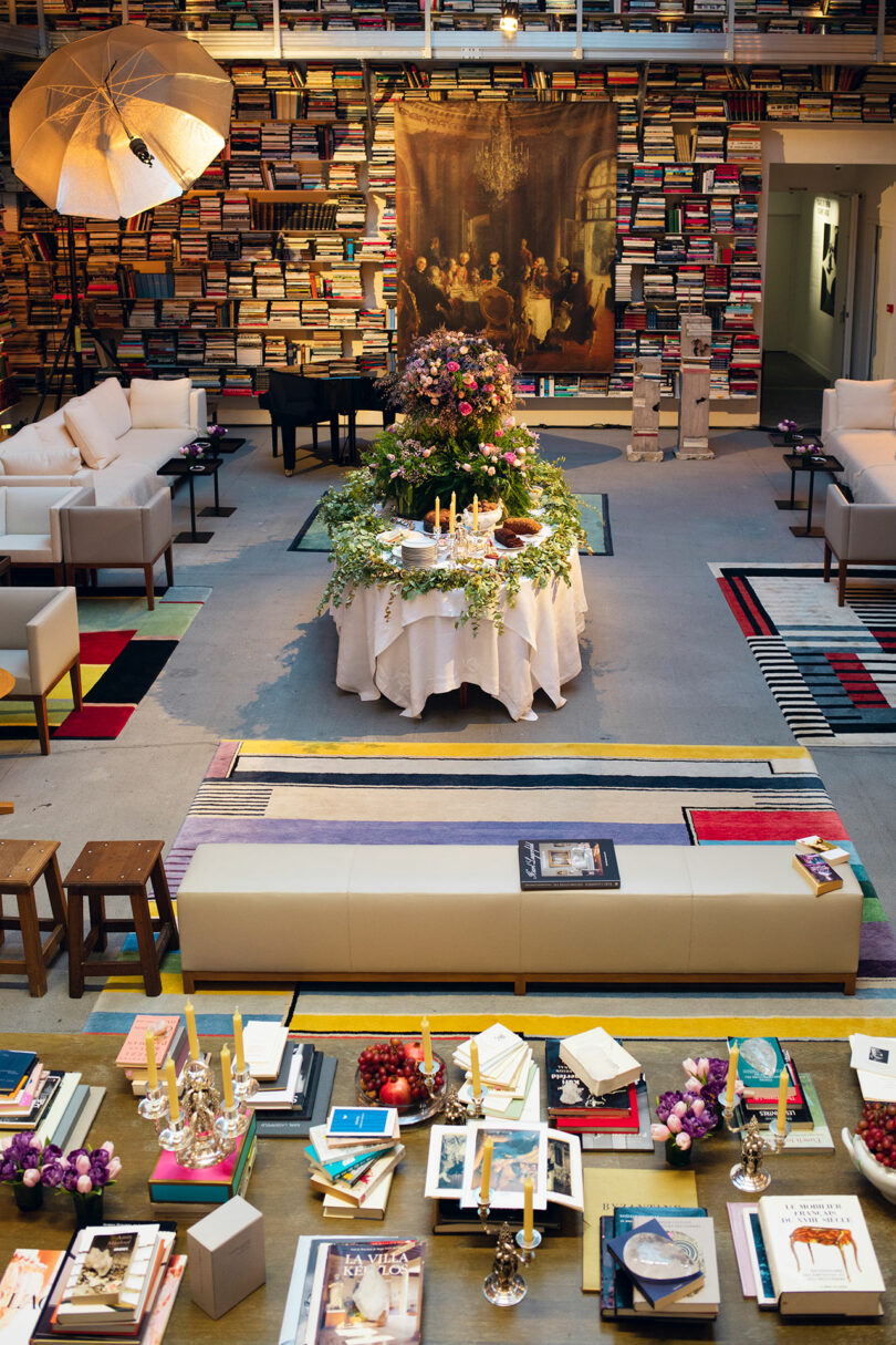 A room filled with stacked books, a table with candles and flowers, sofas, a piano, and decorative tables with books and grapes. An art piece hangs on the wall.