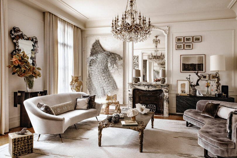 Elegant living room with a combination of modern and vintage decor, featuring a chandelier, plush sofas, a marble fireplace, and large artwork on the walls.